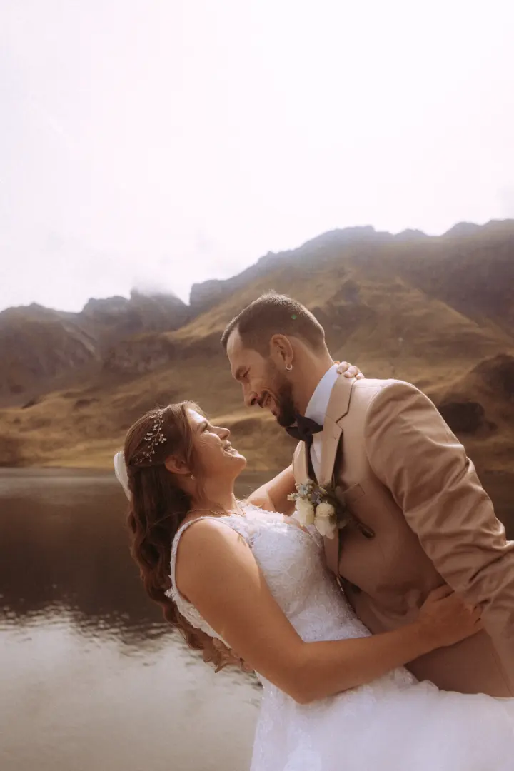 Ein glückliches Brautpaar in einer innigen Pose vor einer majestätischen Bergkulisse an einem See. Der Bräutigam trägt einen beigen Anzug, die Braut ein weißes Spitzenkleid. Kinematischer Hochzeitsfotograf in der Schweiz.
