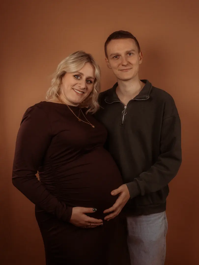 Eine schwangere Frau in einem dunkelroten Kleid und ihr Partner posieren lächelnd für ein professionelles Schwangerschaftsfoto im Fotostudio in Schwerin. Beide legen liebevoll ihre Hände auf den Babybauch vor einem schlichten, braunen Hintergrund.