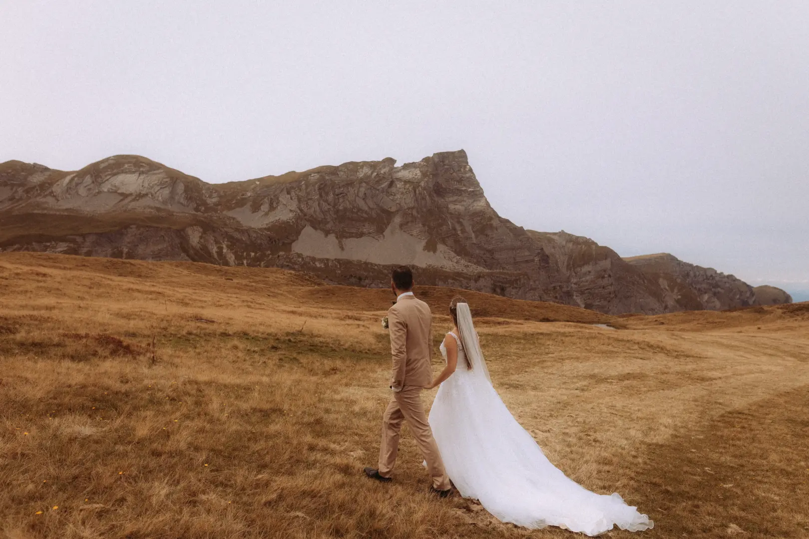 Ein Brautpaar geht Hand in Hand über eine herbstliche Bergwiese in der Schweiz. Die Braut trägt ein weißes Hochzeitskleid mit langem Schleier, der Bräutigam einen beigen Anzug. Im Hintergrund erheben sich majestätische, felsige Berge. Kinematischer Hochzeitsfotograf in der Schweiz.