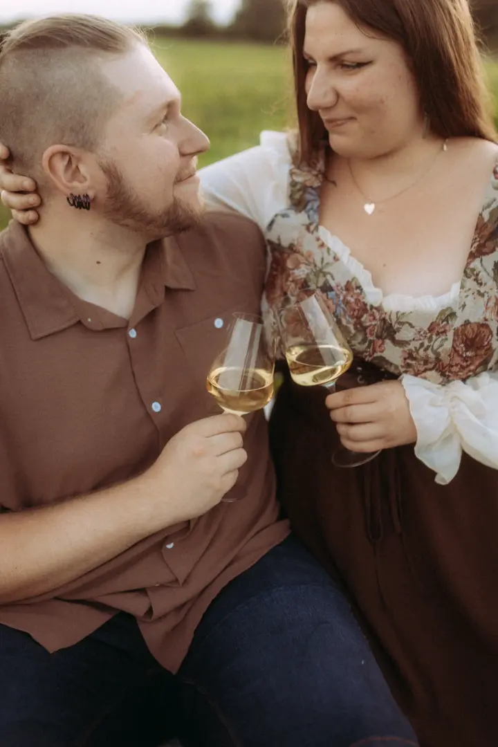 Ein verliebtes Paar stößt bei einem romantischen Picknick im Freien mit Weingläsern an. Die Frau trägt ein florales Oberteil, der Mann ein braunes Hemd. Die Aufnahme fängt eine innige Stimmung ein, perfekt für authentische Pärchenfotos Schwerin.