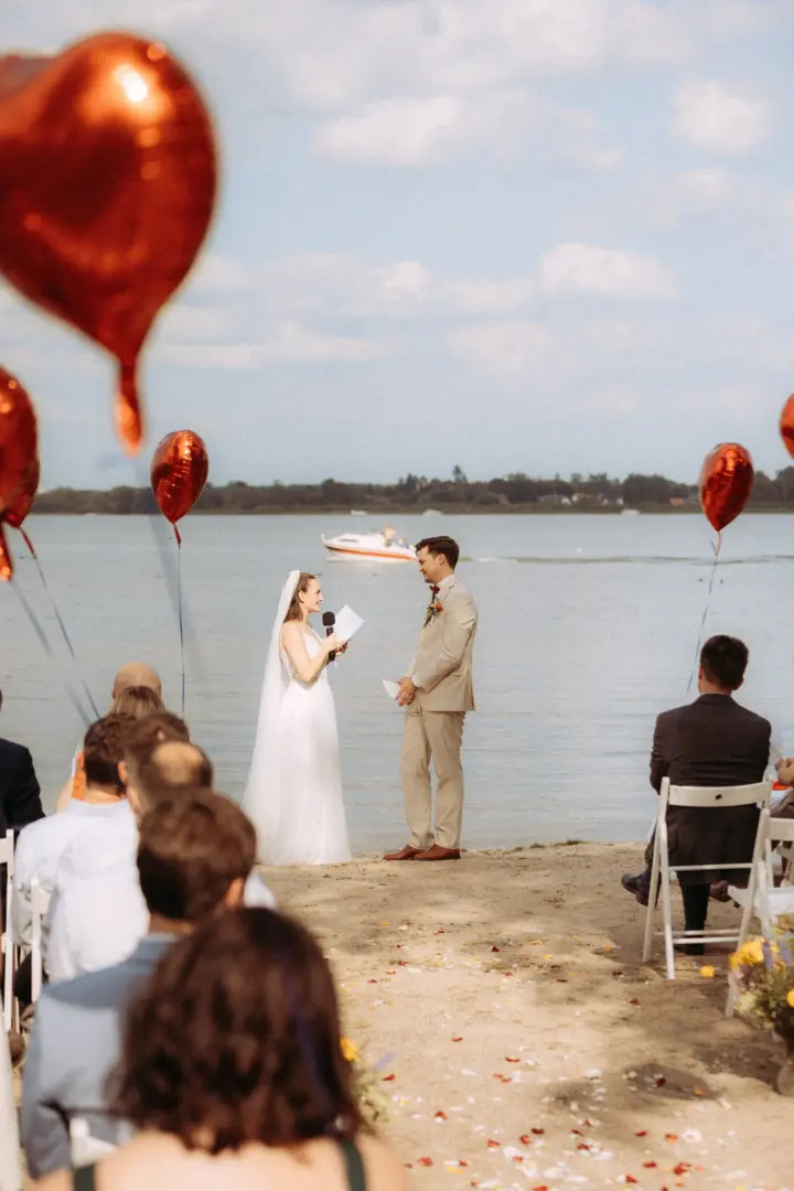 Brautpaar bei der freien Trauung am See, die Braut liest ihr Eheversprechen vor. Im Vordergrund rote Herzballons, im Hintergrund ein vorbeifahrendes Boot. Eine romantische Szene für den Hochzeitsfotograf in Schwerin und MV.