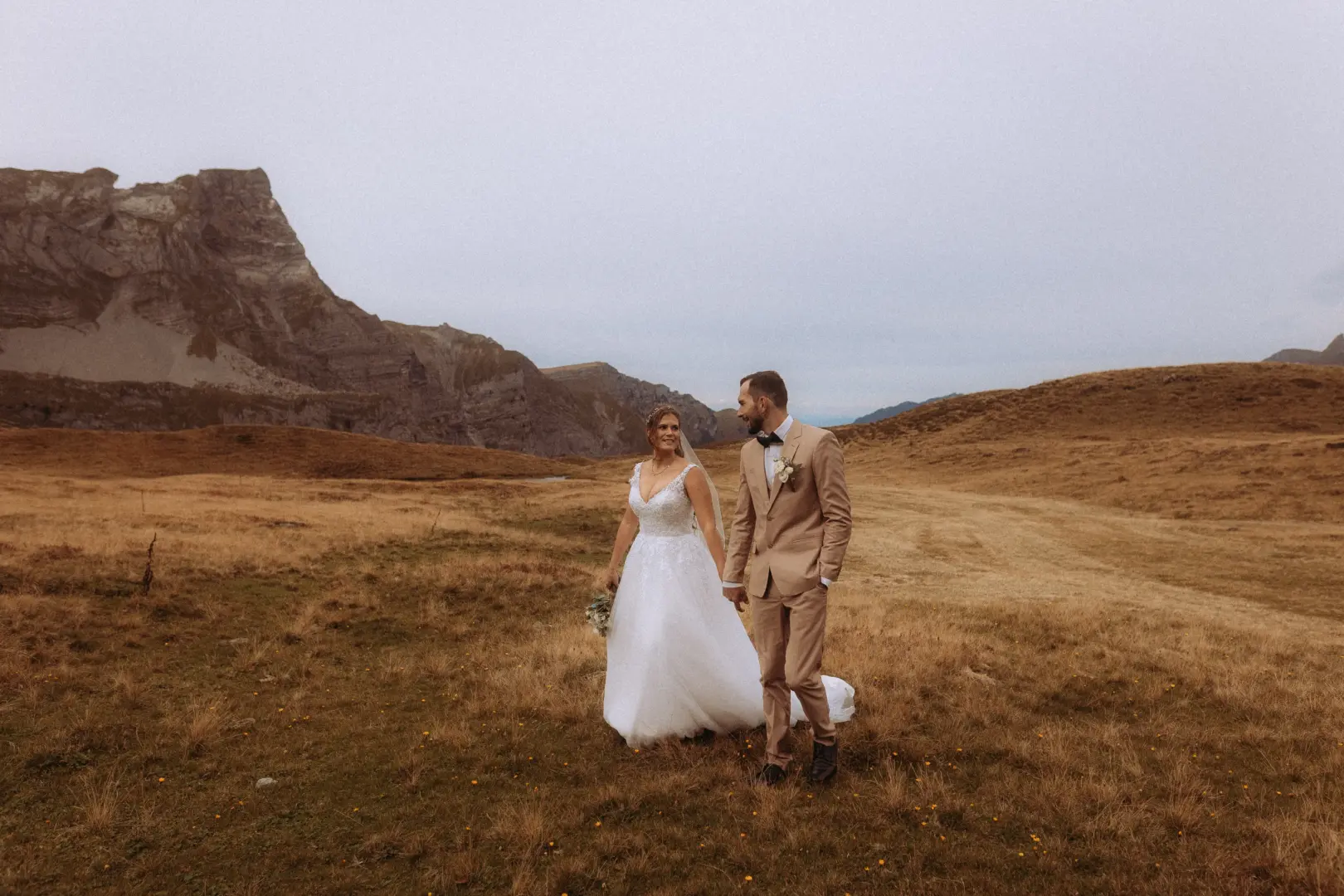 Ein Brautpaar spaziert händchenhaltend durch eine herbstliche Berglandschaft in der Schweiz. Die Braut trägt ein weißes Hochzeitskleid, der Bräutigam einen beigen Anzug. Kinematischer Hochzeitsfotograf in der Schweiz, der diesen romantischen Moment vor imposanter Bergkulisse festhält.