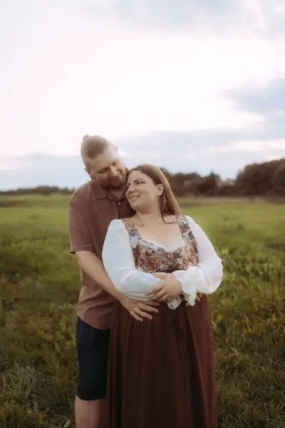 Ein glückliches Paar bei einem romantischen Fotoshooting auf einer weiten Wiese im Abendlicht. Der Mann umarmt die Frau von hinten, beide lächeln sich liebevoll an. Die Szene fängt die Verbundenheit perfekt ein, ideal für authentische Pärchenfotos Schwerin.