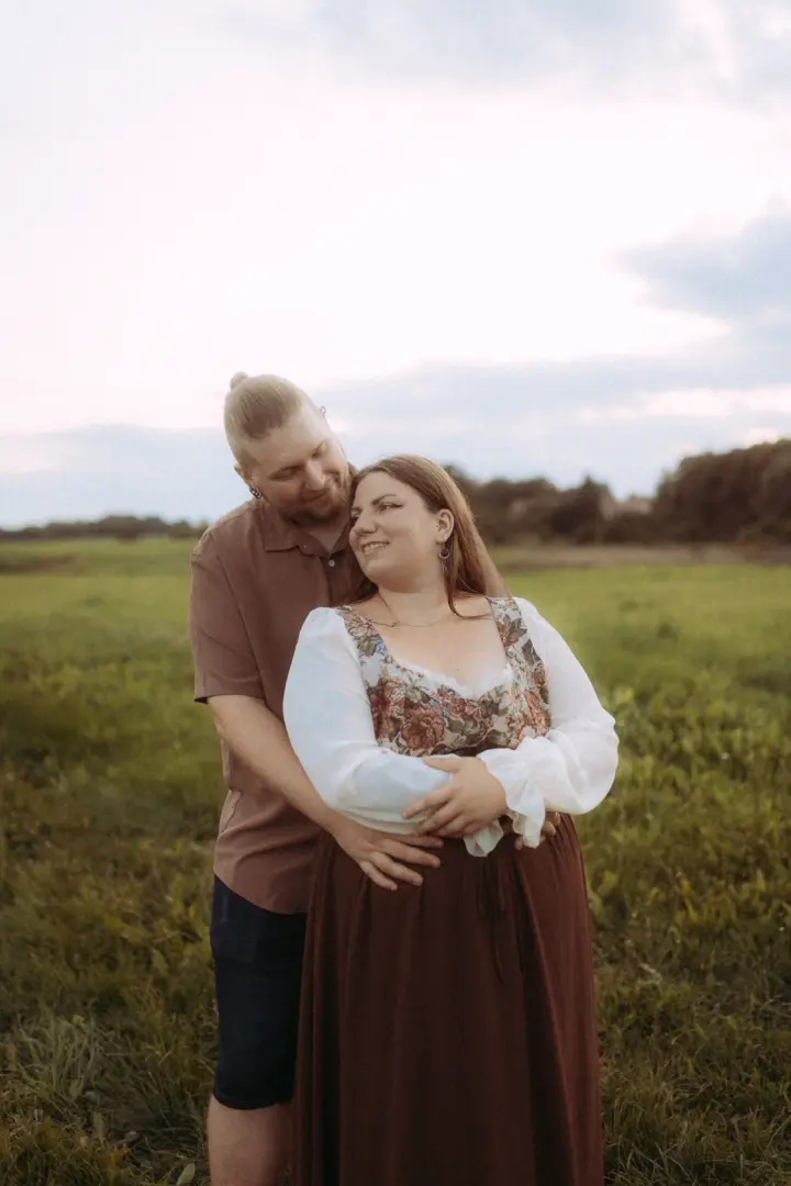 Ein glückliches Paar bei einem romantischen Fotoshooting auf einer weiten Wiese im Abendlicht. Der Mann umarmt die Frau von hinten, beide lächeln sich liebevoll an. Die Szene fängt die Verbundenheit perfekt ein, ideal für authentische Pärchenfotos Schwerin.