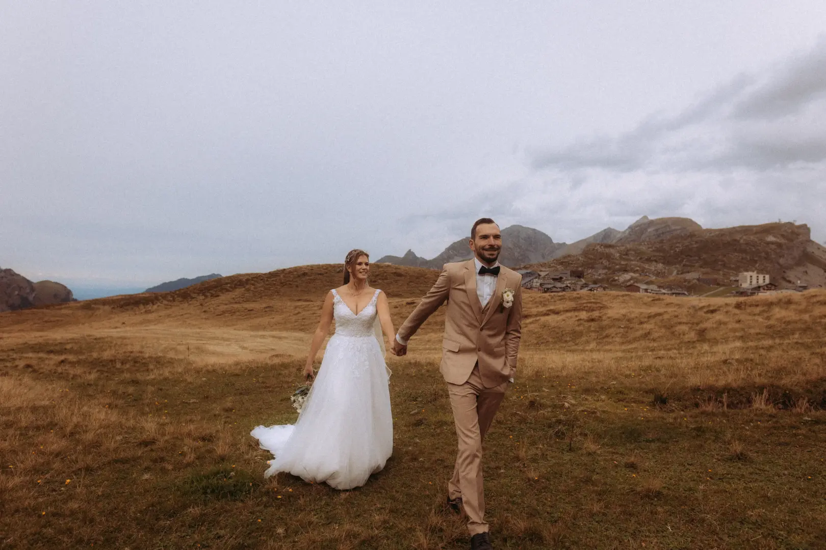 Ein glückliches Brautpaar hält Händchen und läuft über eine weite, herbstliche Bergwiese in den Schweizer Alpen. Der Bräutigam im beigen Anzug und die Braut im weißen Hochzeitskleid strahlen vor einer malerischen Bergkulisse. Kinematischer Hochzeitsfotograf in der Schweiz.