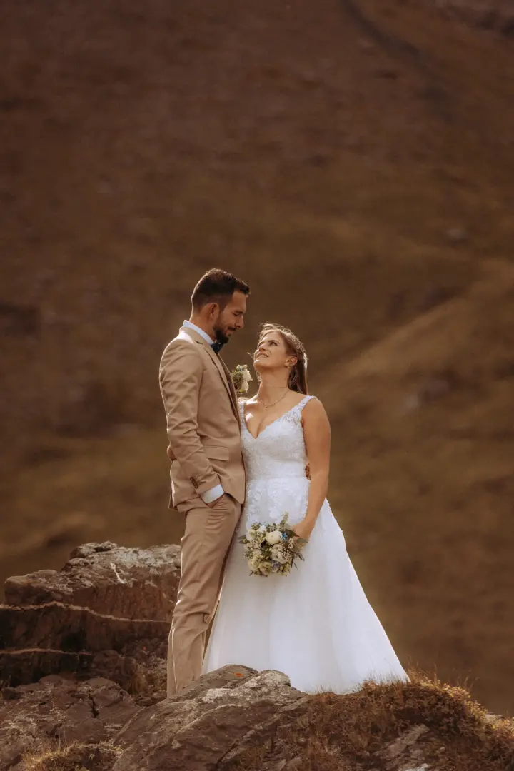 Ein Brautpaar in Hochzeitskleidung steht sich liebevoll auf einem felsigen Berggipfel gegenüber. Die Braut hält einen Blumenstrauß, während der Bräutigam im beigen Anzug sie ansieht. Kinematischer Hochzeitsfotograf in der Schweiz.