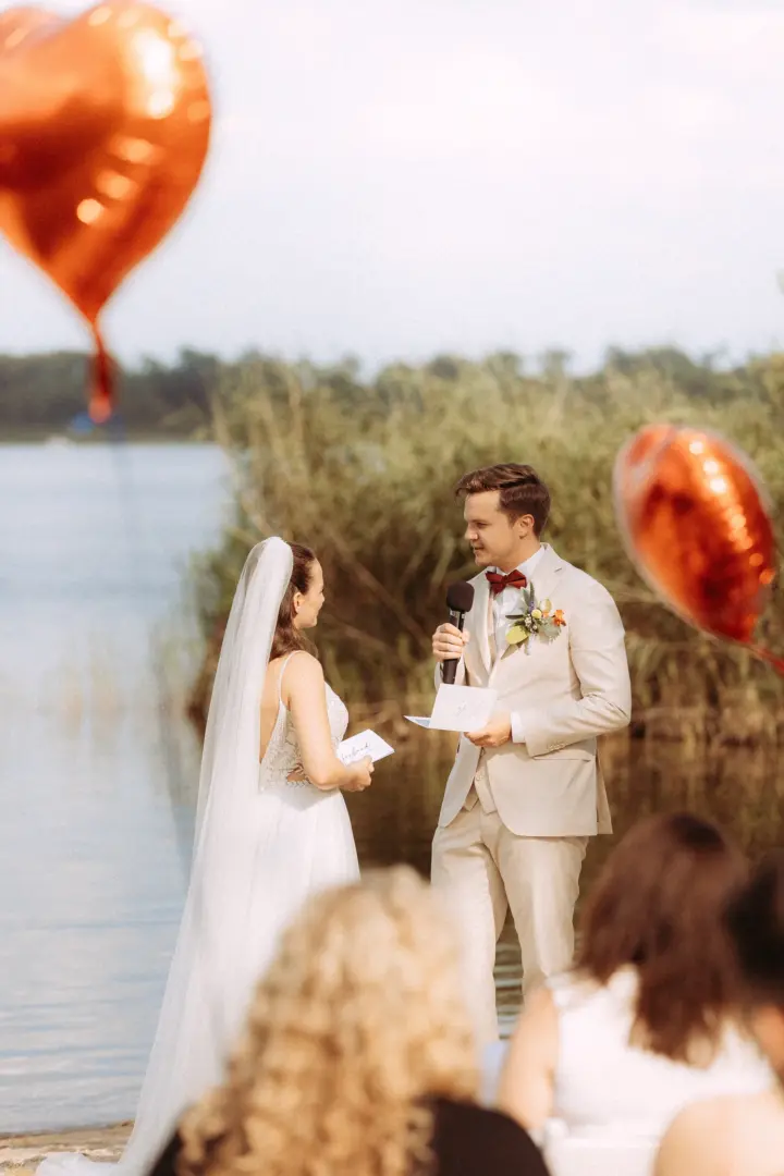 Brautpaar bei der emotionalen Trauung am Seeufer: Der Bräutigam hält eine Rede mit Mikrofon, während rote herzförmige Luftballons im Vordergrund schweben. Stimmungsvolle Aufnahme vom Hochzeitsfotograf in Schwerin und MV.