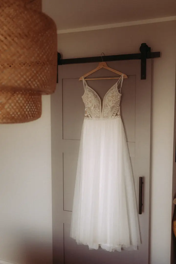 Ein elegantes weißes Brautkleid mit filigraner Spitzenoberseite und fließendem Tüllrock, das an einem Holzkleiderbügel an einer grauen Schiebetür hängt. Eine stimmungsvolle Aufnahme vom Hochzeitsfotograf in Schwerin und MV.