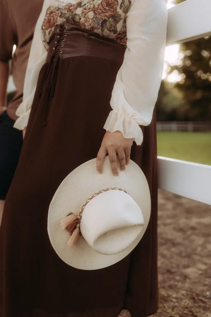 Eine Frau in einem eleganten, bodenlangen braunen Rock mit floralem Oberteil und Korsett hält einen hellen Sommerhut in der Hand. Die Szene, aufgenommen bei einem Fotoshooting für Pärchenfotos in Schwerin, vermittelt eine romantische und natürliche Stimmung vor einem weißen Holzzaun.