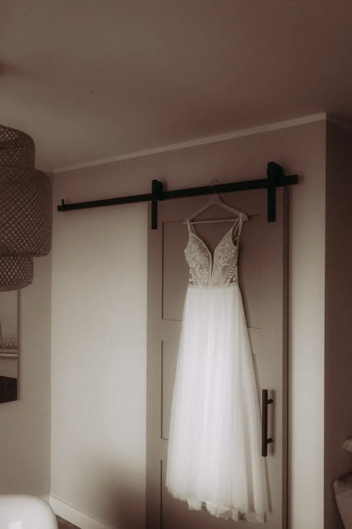 Ein elegantes, weißes Brautkleid mit filigranen Spitzenapplikationen am Oberteil hängt an einem Kleiderbügel an einer modernen Schiebetür, fotografiert von einem Hochzeitsfotograf in Schwerin und MV.