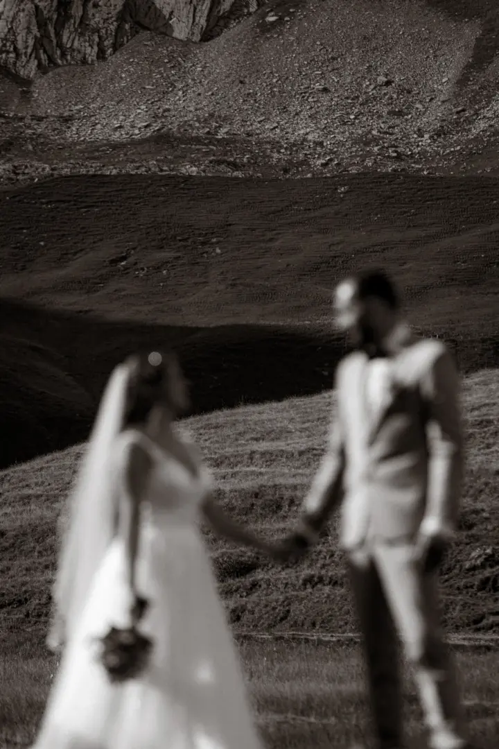 Ein Brautpaar hält sich in einer unscharfen, romantischen Aufnahme vor einer dramatischen Bergkulisse an den Händen. Die Szene wirkt zeitlos und emotional. Kinematischer Hochzeitsfotograf in der Schweiz.