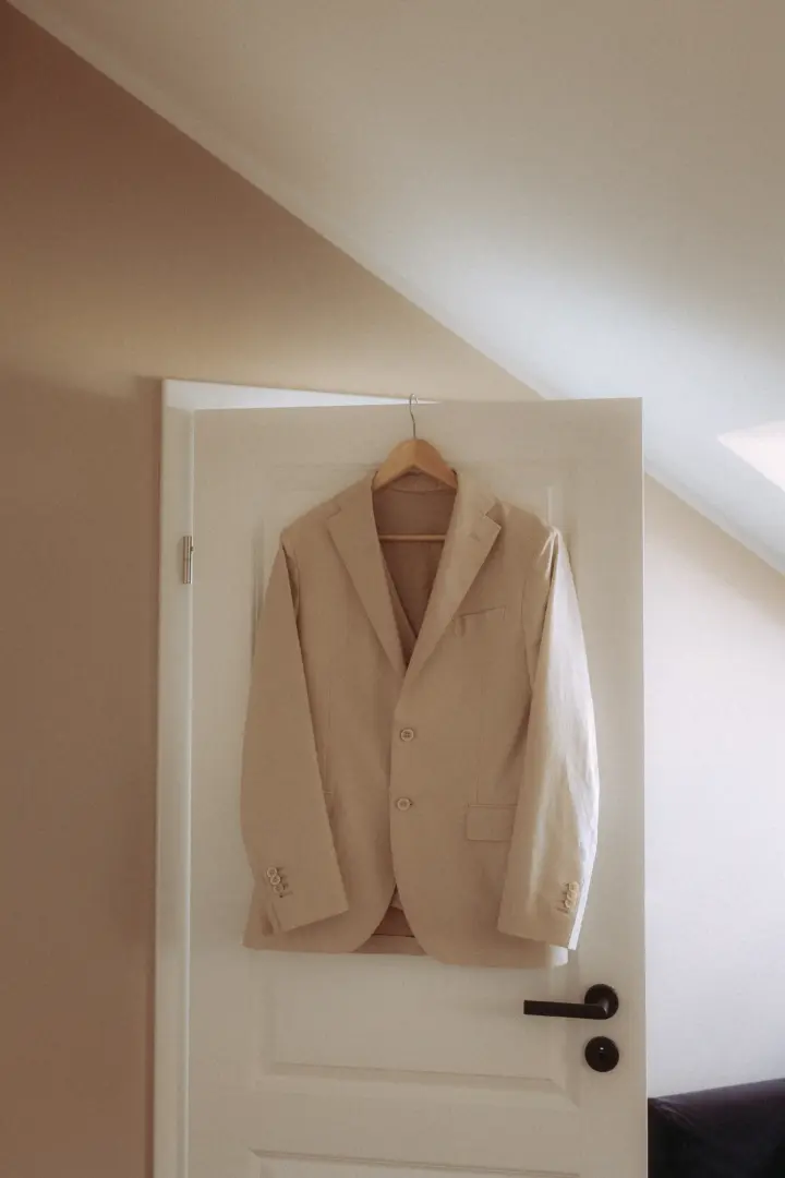 Ein elegantes beiges Sakko hängt an einem Holzkleiderbügel an einer weißen Zimmertür. Das stilvolle Outfit für den Bräutigam ist ein klassisches Detail, das ein Hochzeitsfotograf in Schwerin und MV bei der Vorbereitung festhält.