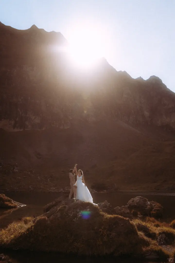 Ein Brautpaar tanzt bei Sonnenuntergang auf einem Felsen vor einer majestätischen Bergkulisse an einem Bergsee. Die dramatische Lichtstimmung unterstreicht diesen romantischen Moment als kinematischer Hochzeitsfotograf in der Schweiz.