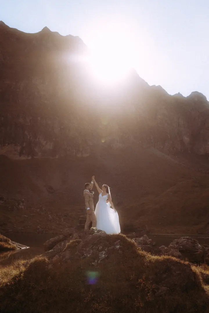Ein Brautpaar tanzt romantisch auf einem Felsen vor einer majestätischen Bergkulisse, während die Sonne hinter den Gipfeln ein warmes Gegenlicht erzeugt. Kinematischer Hochzeitsfotograf in der Schweiz.