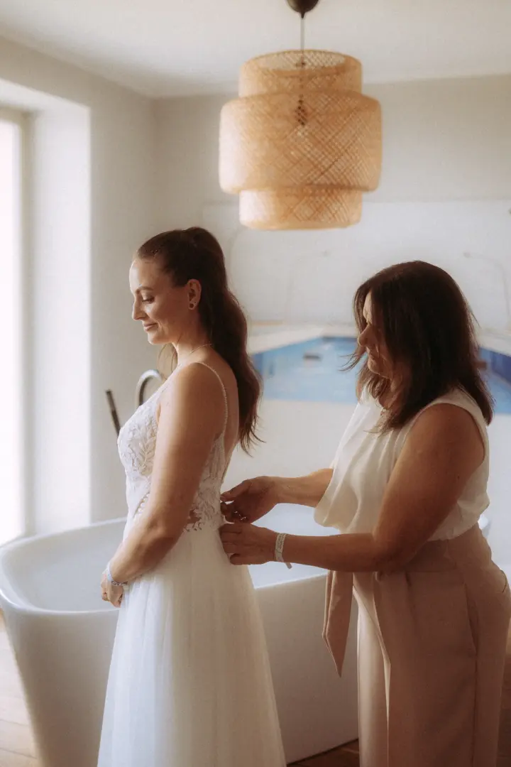 Eine Braut mit offenem Haar trägt ihr weißes Spitzenkleid, während ihr eine Frau beim Schließen des Kleides am Rücken hilft. Das intime Getting-Ready findet in einem hellen, modernen Raum statt. Hochzeitsfotograf in Schwerin und MV.