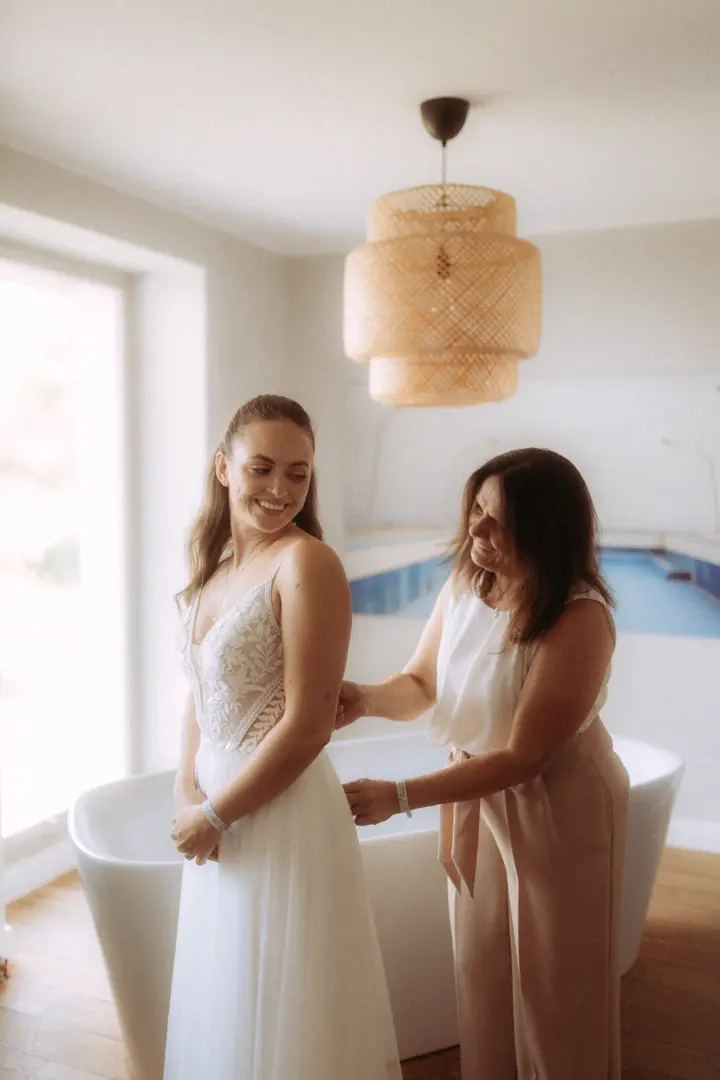 Eine strahlende Braut in einem eleganten weißen Spitzenkleid wird von einer Frau beim Schließen des Kleides unterstützt. Die Szene findet in einem hellen, modernen Raum mit freistehender Badewanne und einer dekorativen Lampe statt, eingefangen vom Hochzeitsfotograf in Schwerin und MV.