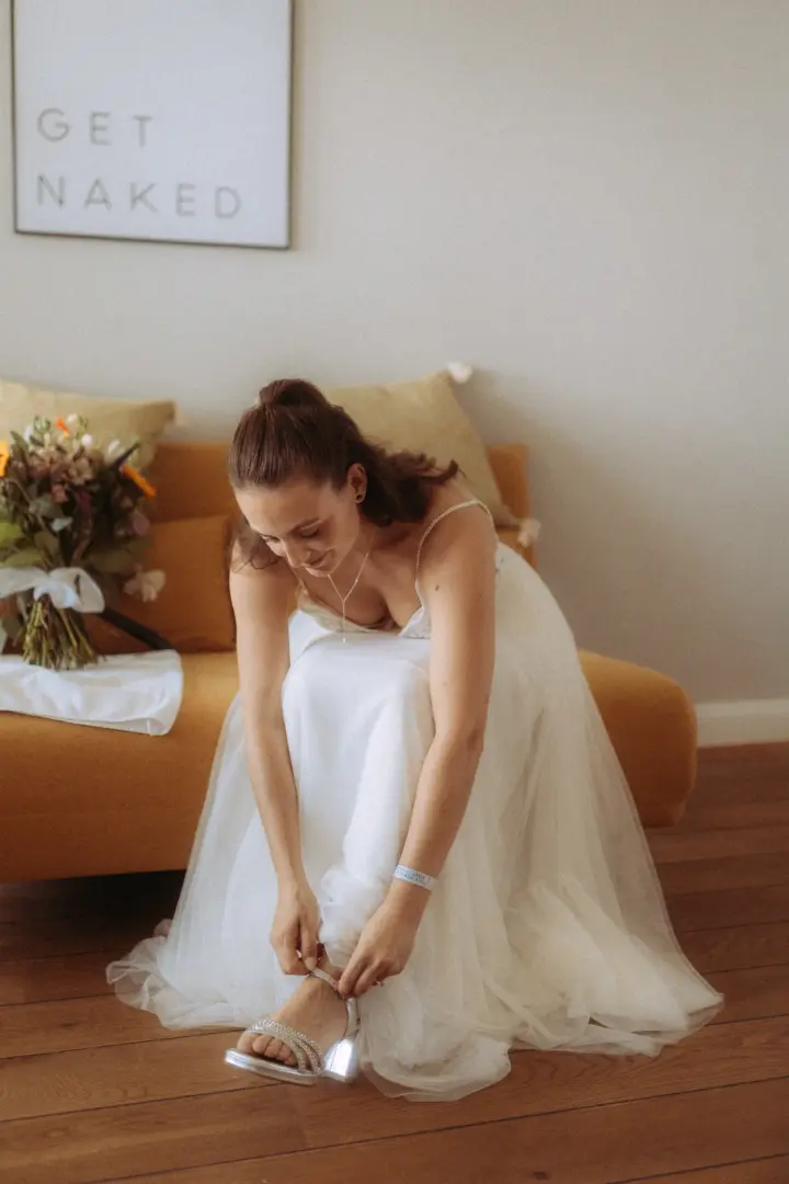 Eine Braut in einem weißen Tüllkleid sitzt auf einem senfgelben Sofa und zieht sich ihre glitzernden Brautschuhe an, während ein Blumenstrauß daneben liegt. Im Hintergrund hängt ein Schild mit der Aufschrift 