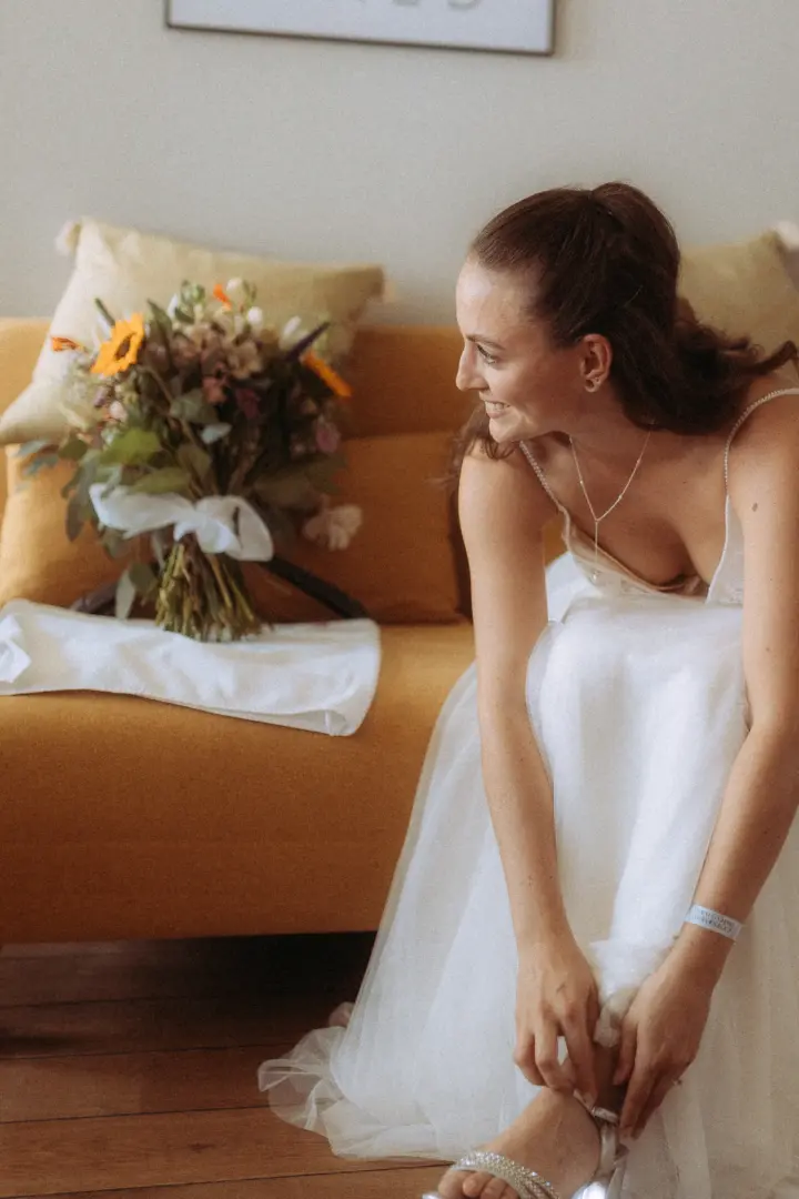 Eine strahlende Braut in einem eleganten weißen Brautkleid zieht sich sitzend auf einem gelben Sofa ihre Schuhe an. Im Hintergrund steht ein bunter Blumenstrauß. Diese Aufnahme zeigt die Vorbereitungen für den großen Tag, festgehalten von Ihrem Hochzeitsfotograf in Schwerin und MV.