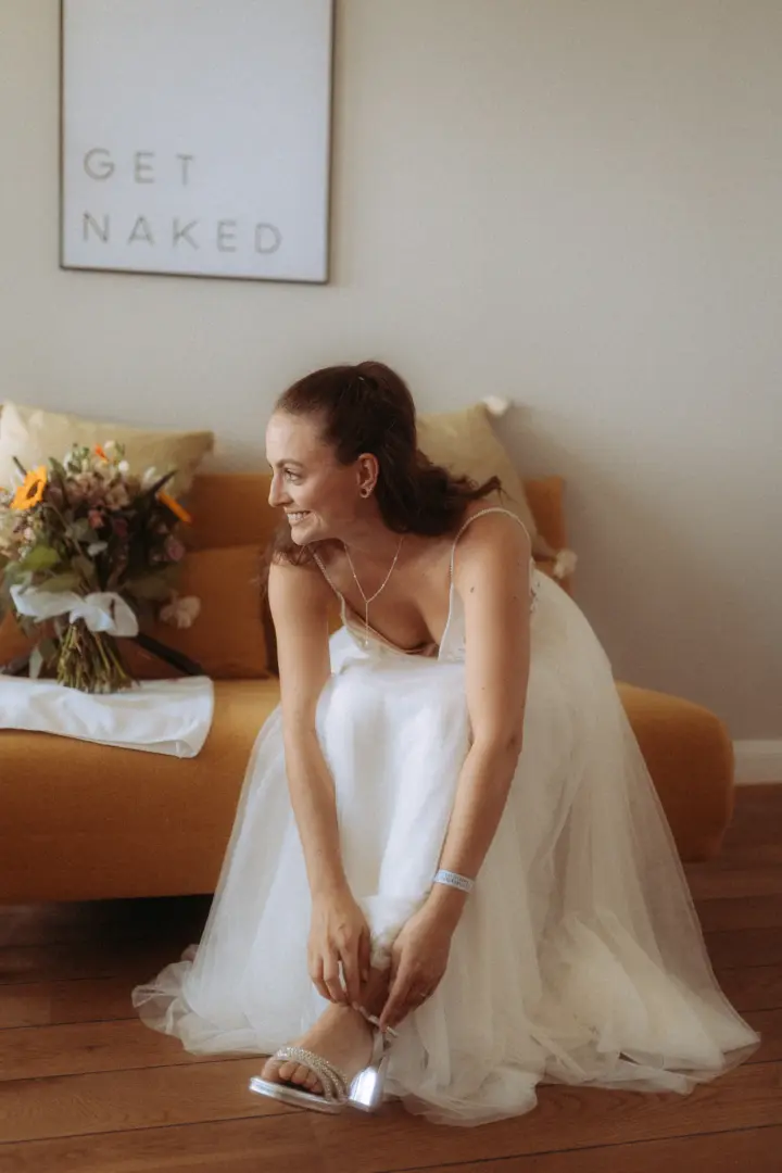 Eine strahlende Braut in einem eleganten weißen Tüllkleid zieht sich sitzend auf einem ockergelben Sofa ihre glitzernden Brautschuhe an. Im Hintergrund hängt ein Bild mit der Aufschrift „GET NAKED“. Authentische Momentaufnahme vom Hochzeitsfotograf in Schwerin und MV.