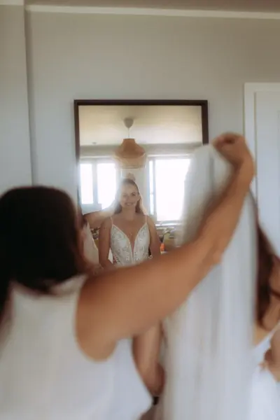 Eine Braut lächelt glücklich in den Spiegel, während ihr Schleier von einer Person im Hintergrund zurechtgerückt wird. Die Szene fängt einen intimen Moment der Hochzeitsvorbereitungen ein, festgehalten von einem Hochzeitsfotograf in Schwerin und MV.