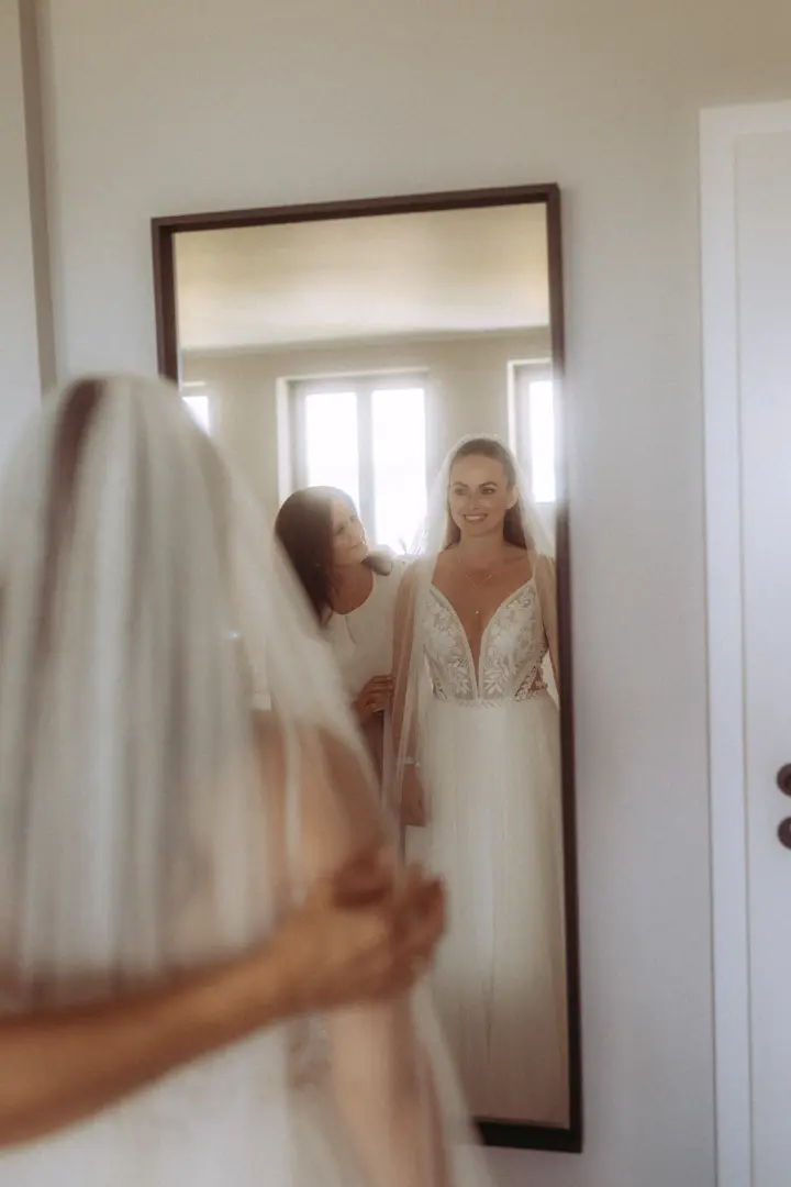Eine Braut mit weißem Spitzenkleid und Schleier betrachtet sich im Spiegel, während eine Frau ihr beim Ankleiden hilft. Diese emotionale Vorbereitung auf den großen Tag wurde von einem Hochzeitsfotograf in Schwerin und MV in einem intimen Moment festgehalten.