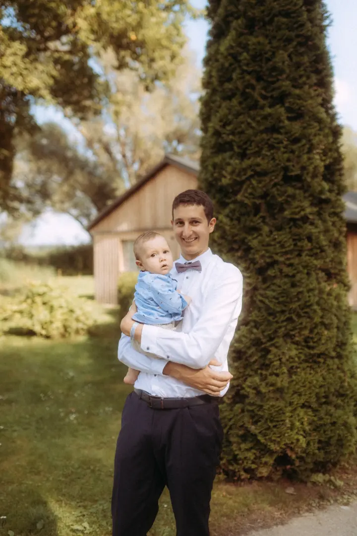 Ein glücklicher Vater im weißen Hemd mit Fliege hält sein Baby im Arm, das ein hellblaues Hemd trägt. Im Hintergrund ist ein sonniger Garten mit einem Holzhaus und einer großen Zypresse zu sehen, festgehalten von einem Fotograf für Familien in Schwerin.