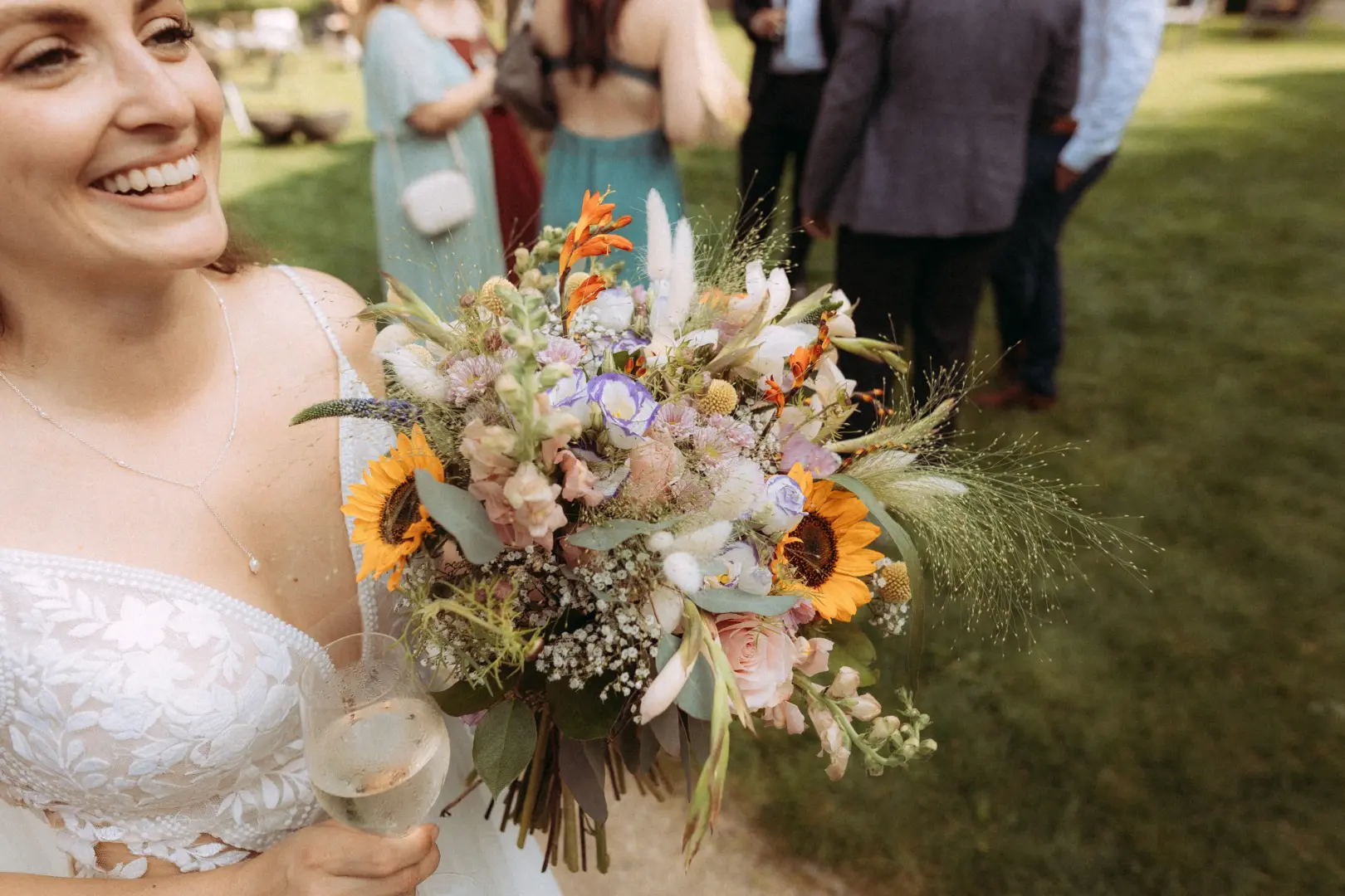 Eine strahlende Braut in einem eleganten Spitzenkleid hält einen bunten Wildblumenstrauß mit Sonnenblumen und ein Glas Sekt in der Hand. Im Hintergrund sind Hochzeitsgäste auf einer grünen Wiese zu sehen. Authentische Aufnahme von einem Hochzeitsfotograf in Schwerin und MV.