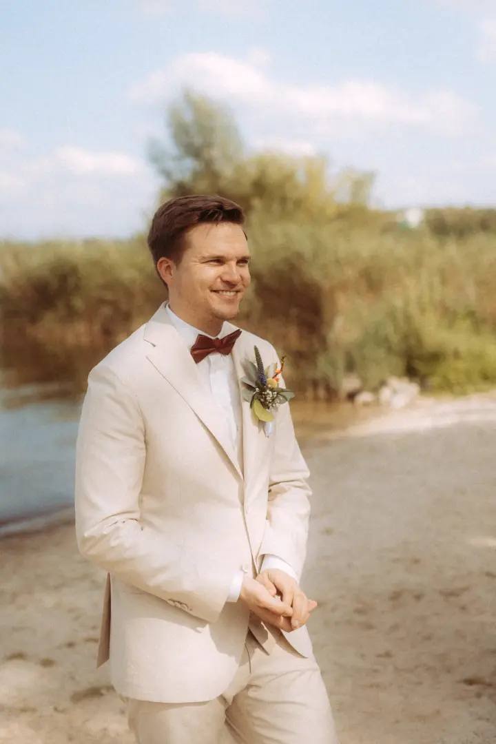 Ein glücklicher Bräutigam in einem beigen Anzug mit dunkelroter Fliege und Blumen-Anstecker lächelt bei einer Strandhochzeit in die Ferne. Diese Aufnahme wurde von einem Hochzeitsfotograf in Schwerin und MV festgehalten.