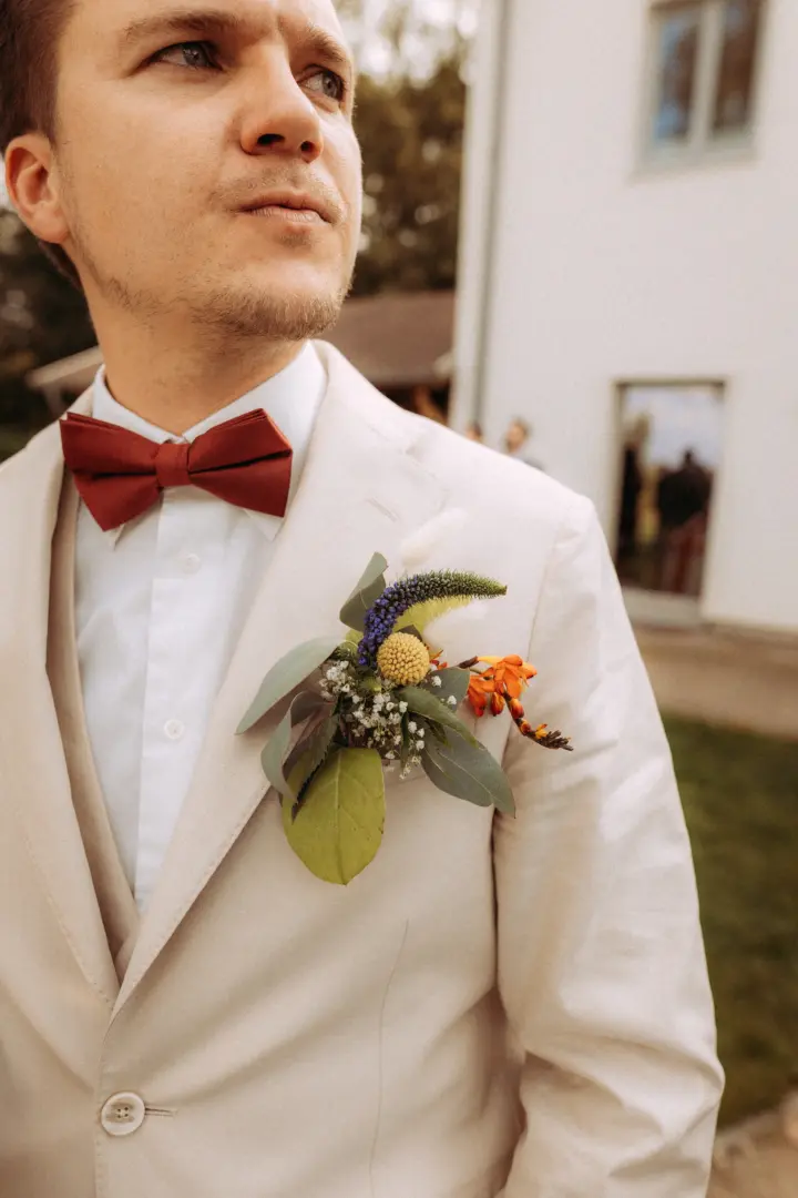 Bräutigam in einem beigen Leinenanzug mit dunkelroter Fliege und einem kunstvollen Ansteckblumen-Bouquet aus Wildblumen, fotografiert von einem Hochzeitsfotograf in Schwerin und MV vor einem hellen Gebäude.