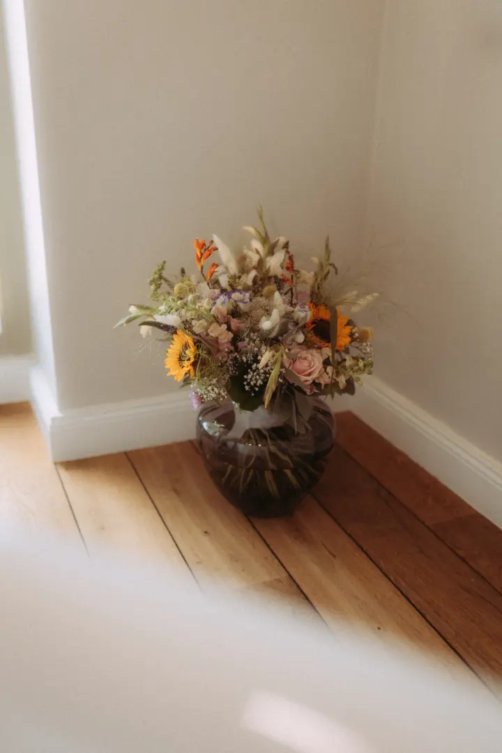 Ein kunstvoller Trockenblumenstrauß mit Sonnenblumen, zarten Rosen und Gräsern steht in einer bauchigen Glasvase in einer Zimmerecke. Das Arrangement sorgt für eine warme, rustikale Atmosphäre und dient als stilvolles Detail für einen Hochzeitsfotograf in Schwerin und MV.