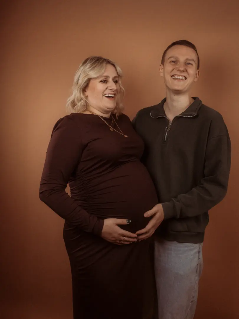 Eine schwangere Frau in einem dunkelroten Kleid und ihr Partner lachen herzlich, während sie gemeinsam ihren Babybauch halten. Das Paar posiert vor einem schlichten, braunen Hintergrund in einem professionellen Fotostudio in Schwerin für authentische Familienfotos.