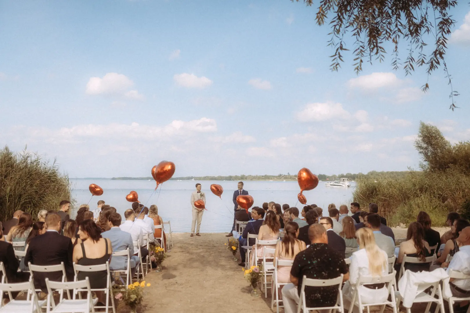 Eine romantische Hochzeit am Seeufer mit Gästen auf weißen Stühlen, die auf das Brautpaar und rote Herz-Luftballons blicken. Ein Hochzeitsfotograf in Schwerin und MV hat diesen besonderen Moment unter blauem Himmel und in idyllischer Natur festgehalten.