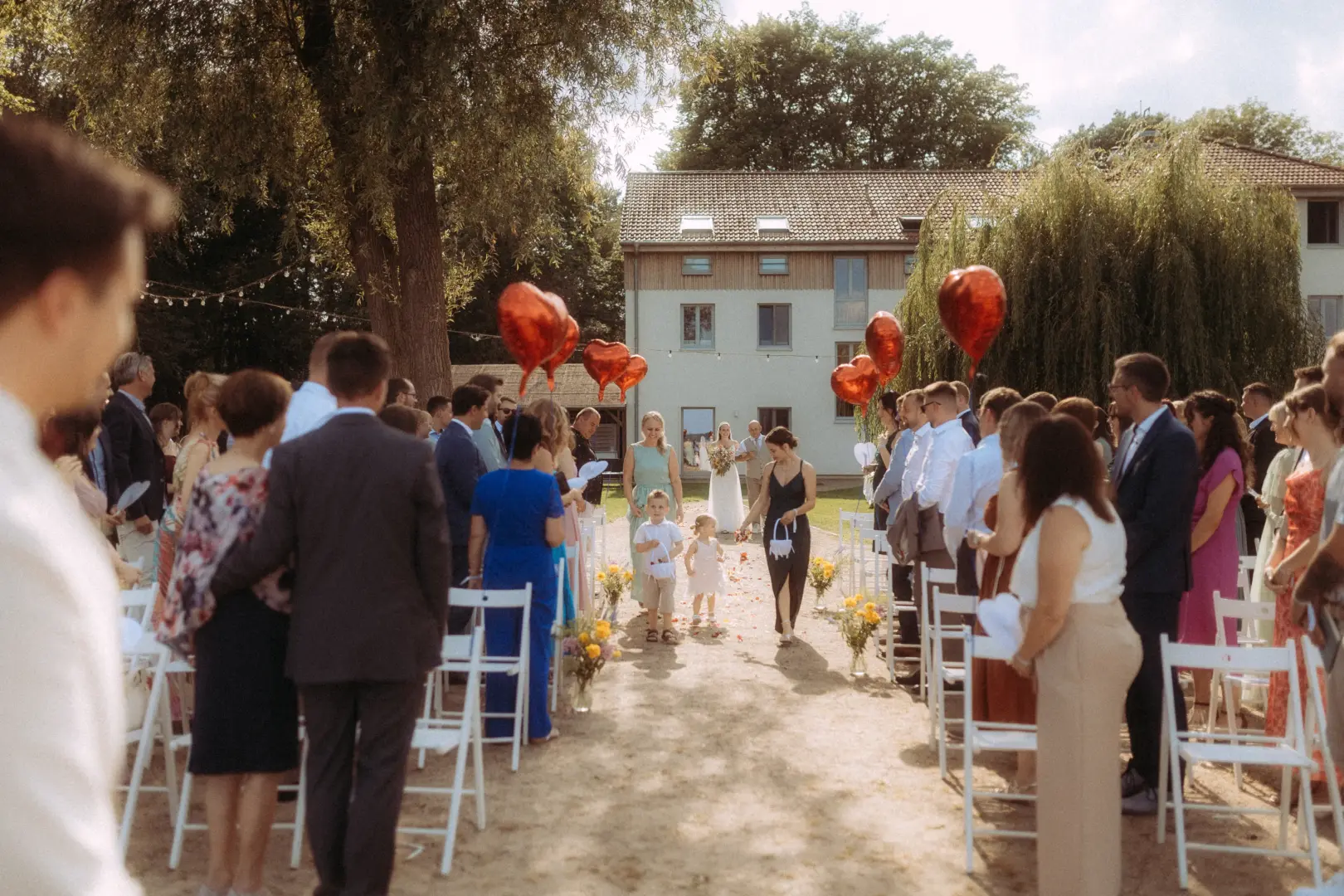 Ein Blumenmädchen und ein Ringträger schreiten bei einer sommerlichen Gartenhochzeit den Gang entlang, während Gäste zuschauen. Rote herzförmige Luftballons säumen den Weg vor einem historischen Gebäude. Aufnahme von einem Hochzeitsfotograf in Schwerin und MV.