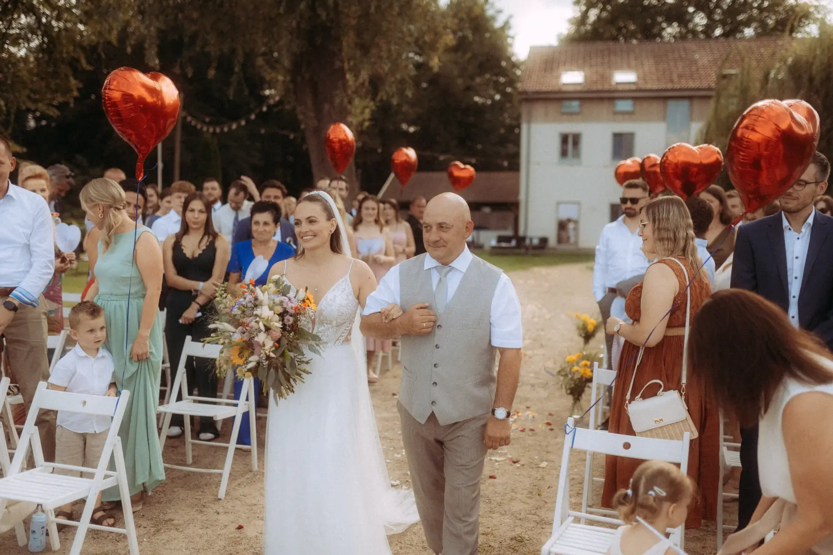 Eine Braut in einem weißen Brautkleid mit Blumenstrauß schreitet an der Seite ihres Vaters den Gang entlang, während rote herzförmige Luftballons im Hintergrund schweben – festgehalten von einem Hochzeitsfotograf in Schwerin und MV.