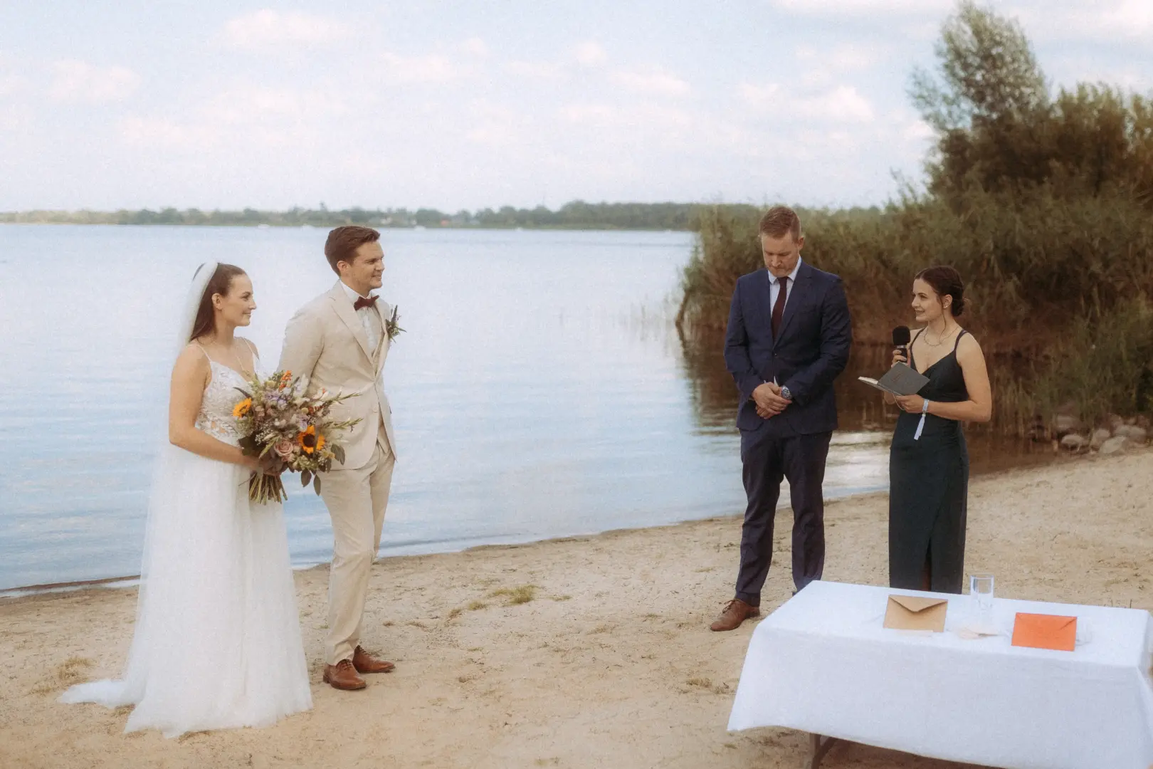 Brautpaar bei einer freien Trauung am Seeufer, begleitet von einem Hochzeitsfotograf in Schwerin und MV. Die Braut im weißen Kleid mit Blumenstrauß und der Bräutigam im hellen Anzug stehen sich gegenüber, während eine Rednerin die Zeremonie am Strand leitet.