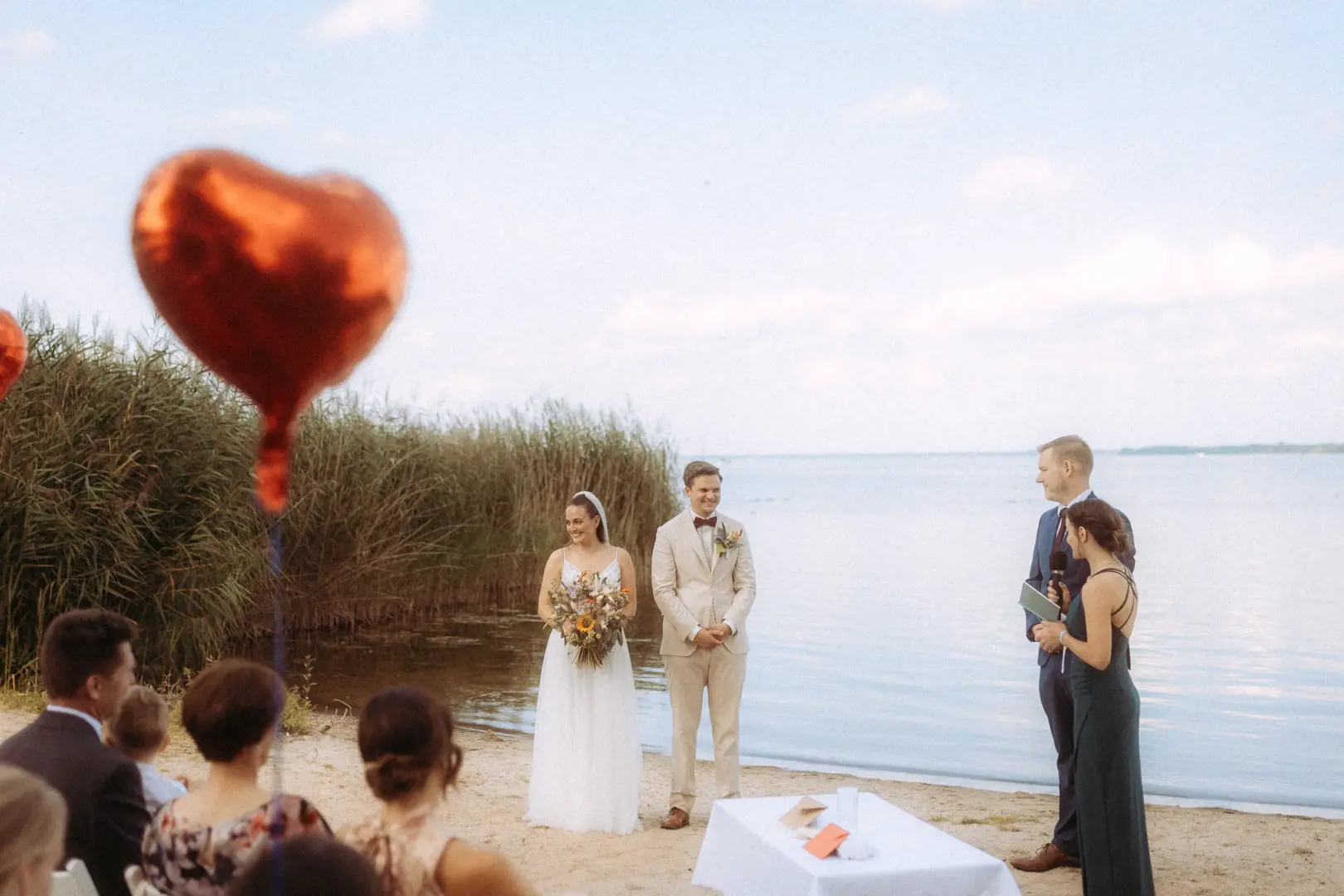 Ein Brautpaar steht während der Trauung am Seeufer, im Vordergrund schwebt ein roter Herzballon. Die Zeremonie wird von einem Trauredner begleitet, während die Gäste im Sand sitzen. Eine stimmungsvolle Aufnahme von Ihrem Hochzeitsfotograf Müritz und MV.