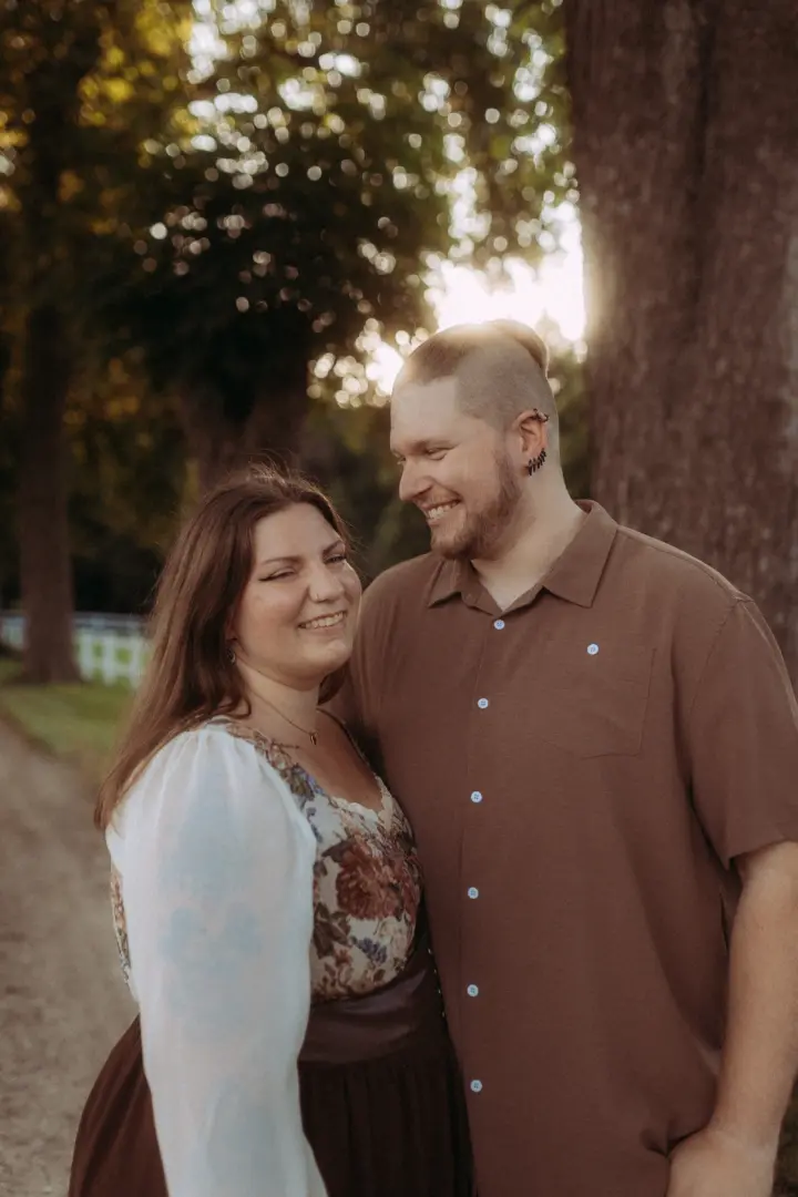 Ein glückliches Paar bei einem romantischen Fotoshooting im Freien während des goldenen Lichts. Der Mann mit Bart und Undercut lächelt seine Partnerin liebevoll an, die ein florales Kleid trägt. Authentische Pärchenfotos Schwerin für unvergessliche Momente.