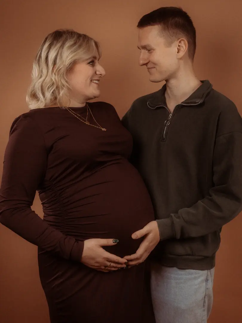 Ein werdendes Elternpaar in einem Fotostudio in Schwerin bei einem liebevollen Schwangerschaftsshooting. Die Frau trägt ein dunkelrotes Kleid und der Mann einen grauen Pullover, während beide gemeinsam ihren Babybauch halten und sich glücklich anlächeln vor einem schlichten, braunen Hintergrund.