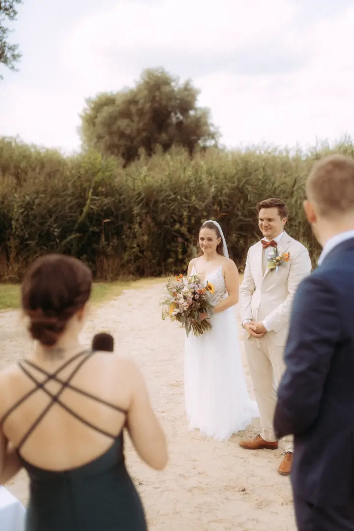 Ein Brautpaar steht sich während einer freien Trauung im Freien gegenüber, während ein Hochzeitsfotograf in Schwerin und MV den Moment festhält. Die Braut trägt ein weißes Spitzenkleid und hält einen bunten Blumenstrauß, der Bräutigam einen hellen Anzug mit Fliege vor einer Naturkulisse.
