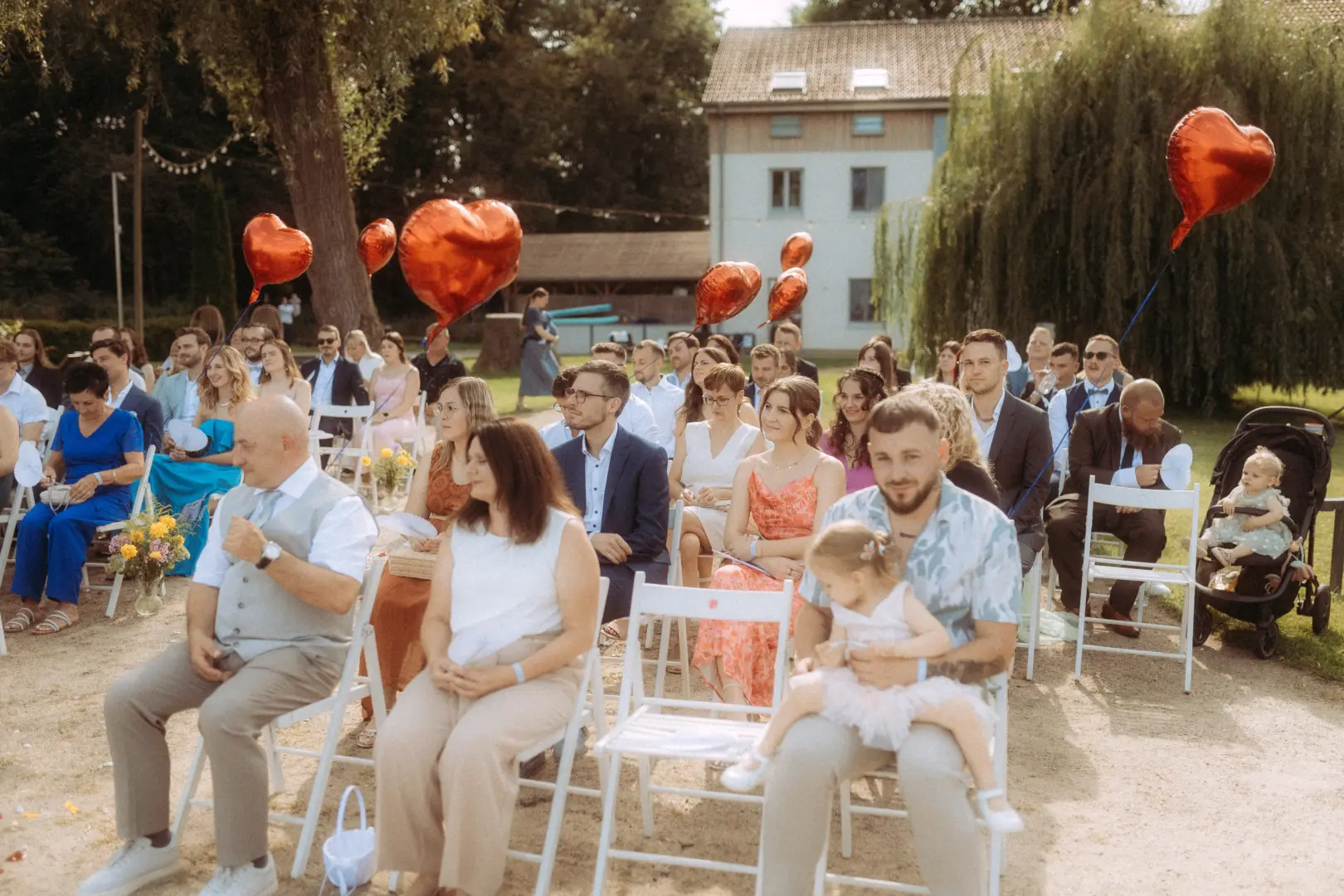 Hochzeitsgäste sitzen bei einer sonnigen Outdoor-Trauung auf weißen Stühlen im Garten. Rote herzförmige Luftballons schweben im Hintergrund, während ein Hochzeitsfotograf in Schwerin und MV die emotionale Zeremonie in einer entspannten, sommerlichen Atmosphäre festhält.