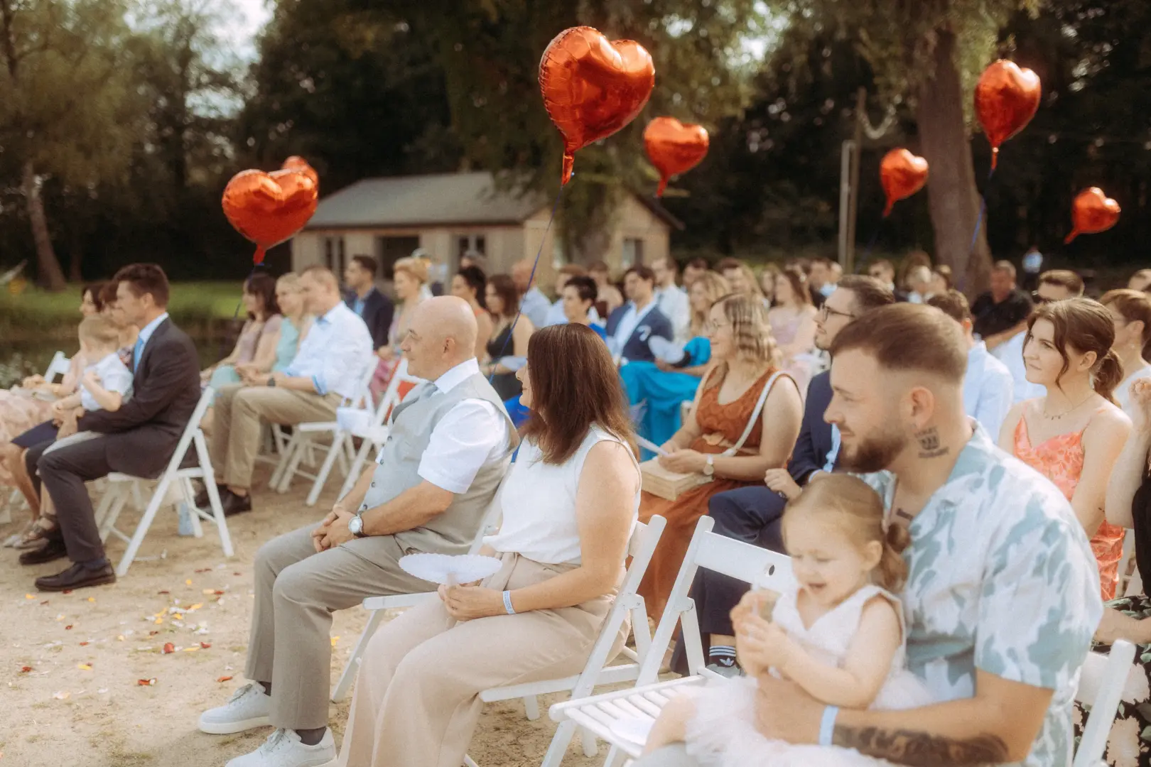 Gäste bei einer sommerlichen Outdoor-Hochzeit sitzen auf weißen Stühlen am Seeufer, geschmückt mit roten herzförmigen Luftballons. Die festliche Atmosphäre wird von einem Hochzeitsfotograf in Schwerin und MV stimmungsvoll eingefangen.