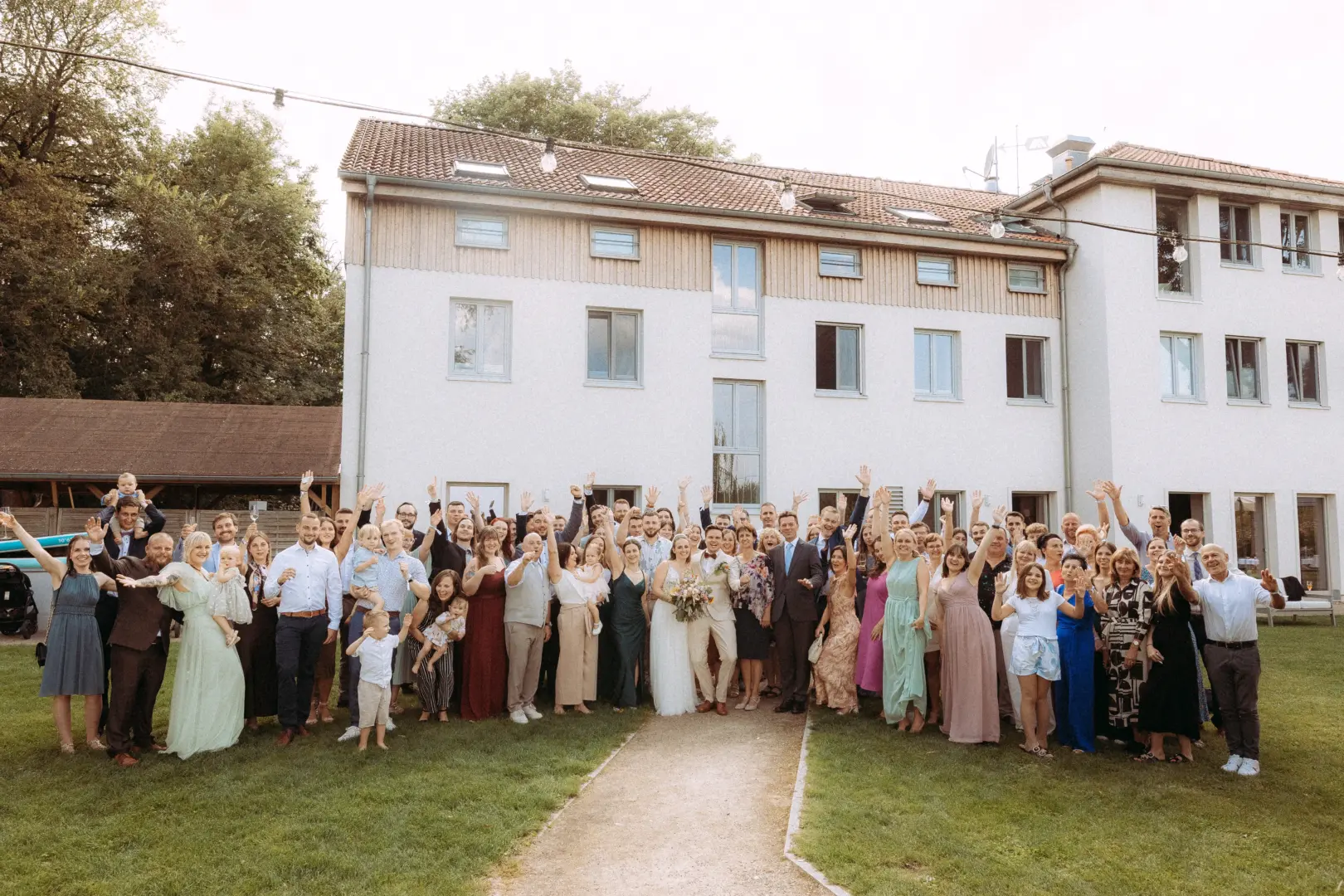 Eine große Hochzeitsgesellschaft feiert im Freien vor einem hellen Gebäude. Das Brautpaar steht in der Mitte, umgeben von glücklichen Gästen, die ihre Arme jubelnd in die Luft strecken. Ein professioneller Hochzeitsfotograf in Schwerin und MV hat diesen freudigen Moment eingefangen.