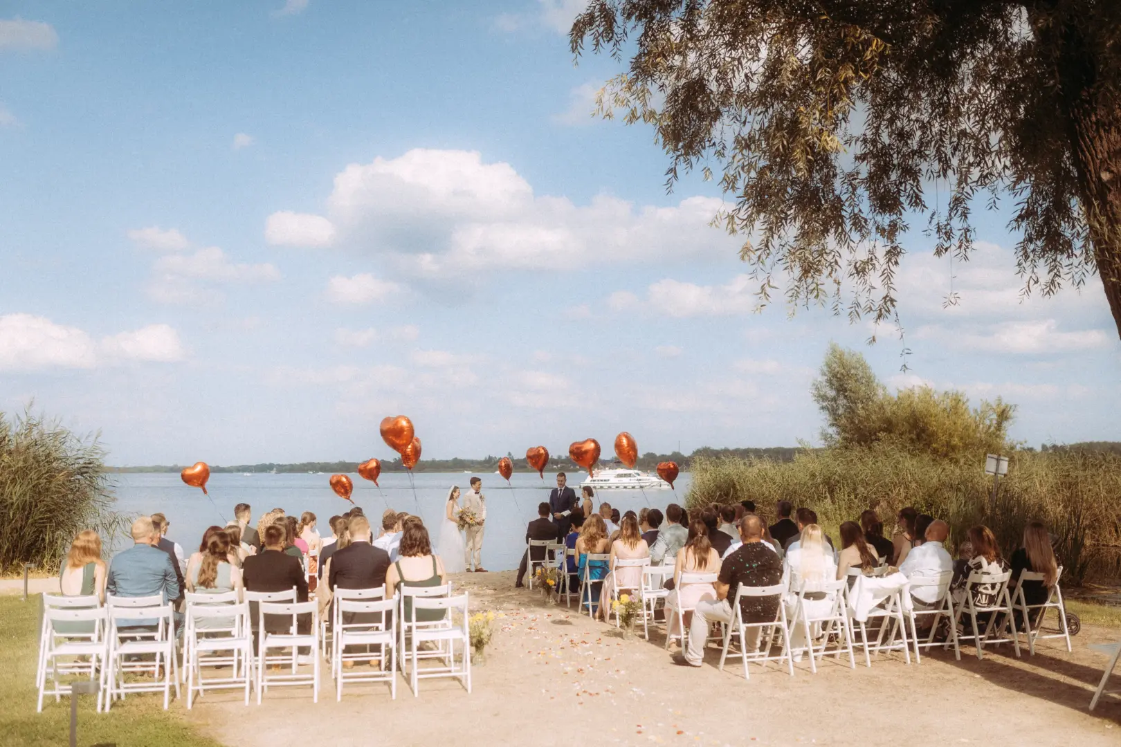 Eine romantische Trauung am Seeufer mit Gästen auf weißen Stühlen, dekoriert mit roten Herzballons. Brautpaar steht vor dem Wasser, im Hintergrund ist ein Schiff zu sehen. Professionelle Aufnahme von einem Hochzeitsfotograf in Schwerin und MV.