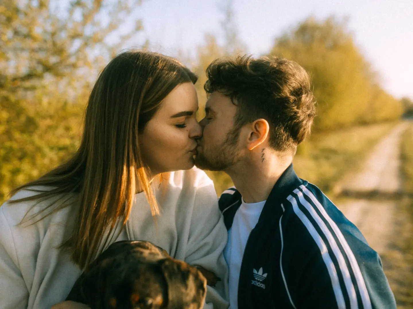 Ein junges Paar küsst sich zärtlich bei warmem Sonnenlicht auf einem ländlichen Weg, während sie gemeinsam ihren Hund halten. Ein romantischer Moment für Pärchenfotos in Schwerin, der die tiefe Verbundenheit in einer natürlichen Umgebung einfängt.