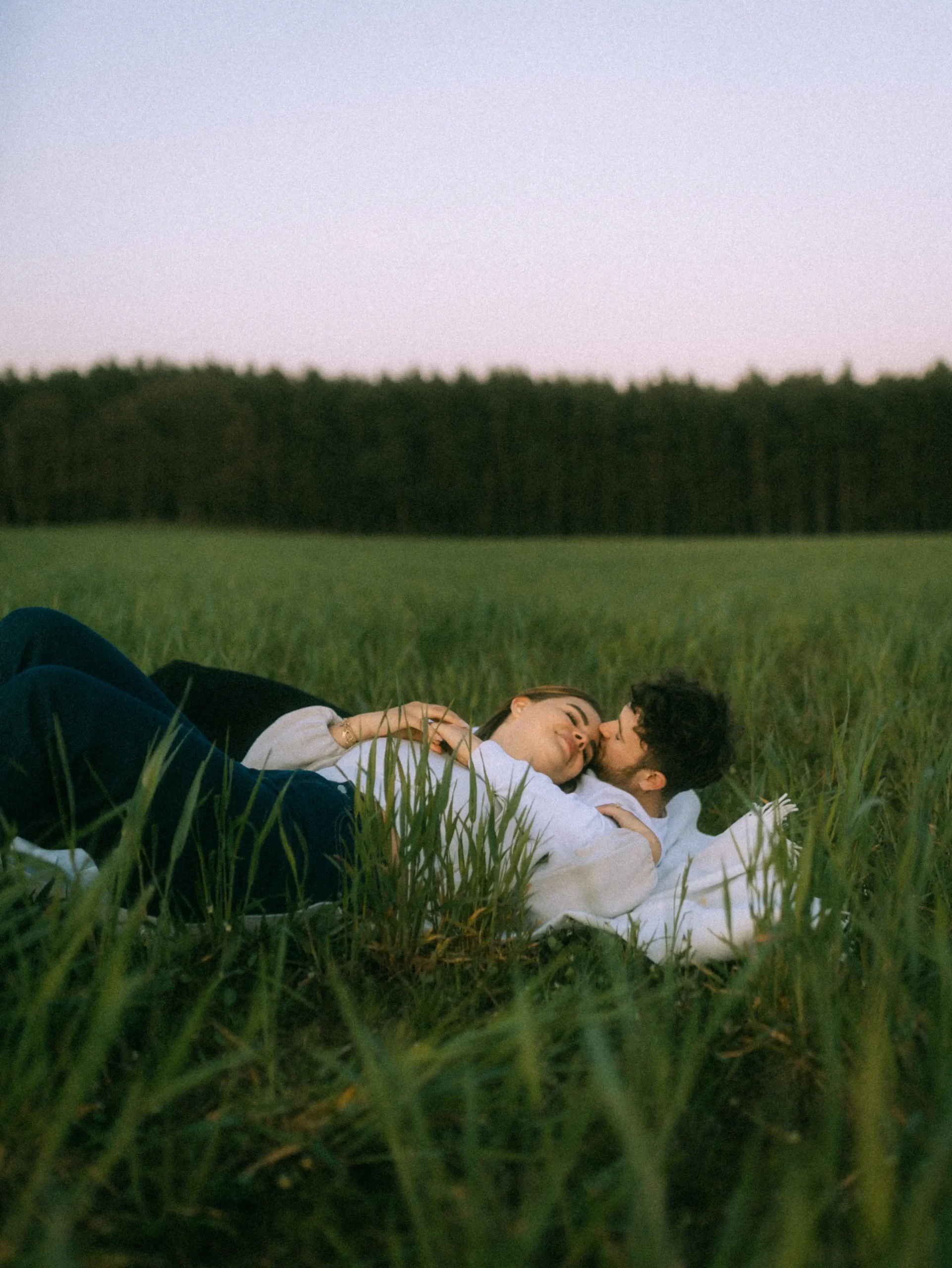 Ein junges Paar liegt eng umschlungen auf einer grünen Wiese im Abendlicht. Der Mann küsst die Frau sanft auf die Wange, während sie die Augen schließt. Ein romantisches Bild einer Lovestory, fotografiert von einem Fotografen für Pärchen in Schwerin und MV. Ein junges Paar liegt eng umschlungen auf einer grünen Wiese im Abendlicht. Der Mann küsst die Frau sanft auf die Wange, während sie die Augen schließt. Ein romantisches Bild einer Lovestory, fotografiert von einem Fotografen für Pärchen in Schwerin und MV.