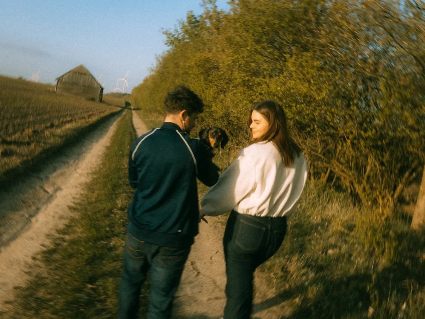 Ein Paar bei einem entspannten Spaziergang auf einem ländlichen Feldweg mit ihrem Dackel, eingefangen während der goldenen Stunde für authentische Pärchenfotos Schwerin. Die warme Abendsonne beleuchtet die beiden und ihre natürliche Verbundenheit in der Natur.