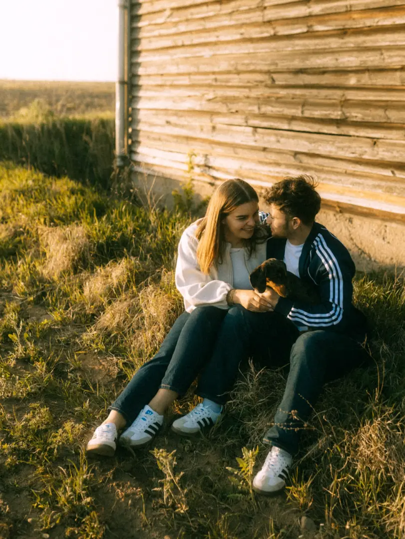 Ein junges Paar sitzt in der warmen Abendsonne vor einer rustikalen Holzwand im Gras und hält liebevoll einen kleinen Dackel im Arm. Die beiden lachen sich glücklich an, während sie entspannte Freizeitkleidung tragen. Authentische Pärchenfotos Schwerin in natürlicher Atmosphäre.