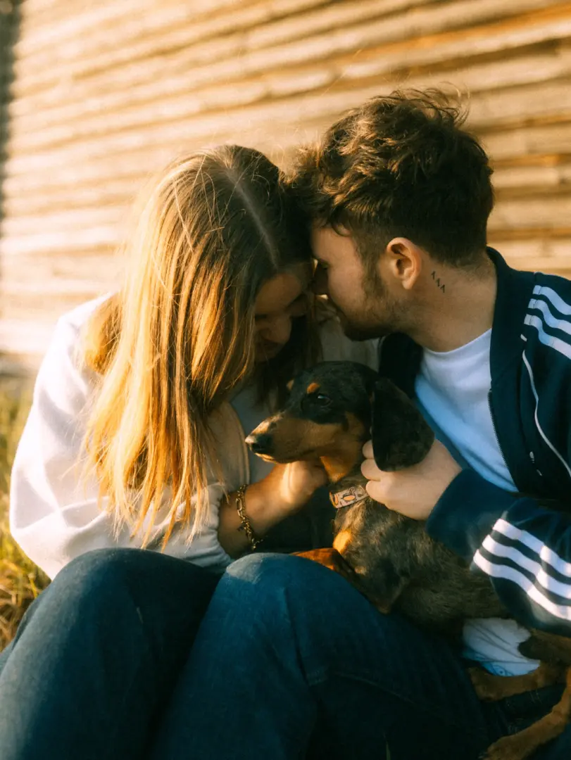 Ein junges Paar sitzt eng beieinander vor einer hölzernen Wand im warmen Licht der Abendsonne und hält liebevoll einen kleinen Dackel im Arm. Die romantische Stimmung unterstreicht die Arbeit als Lovestory Fotograf in Schwerin und MV.