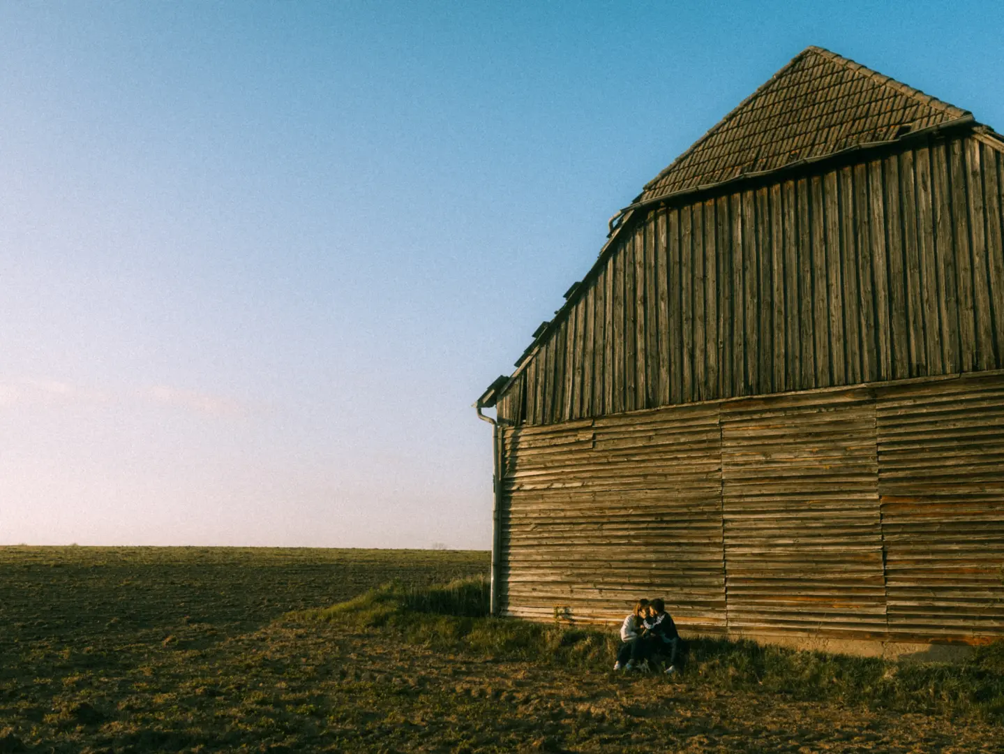 Ein verliebtes Paar sitzt eng beieinander im Gras vor einer rustikalen, alten Holzhütte inmitten eines weiten Feldes während der goldenen Stunde. Romantische Pärchenfotos Schwerin, die die intime Stimmung und die ländliche Kulisse einfangen.