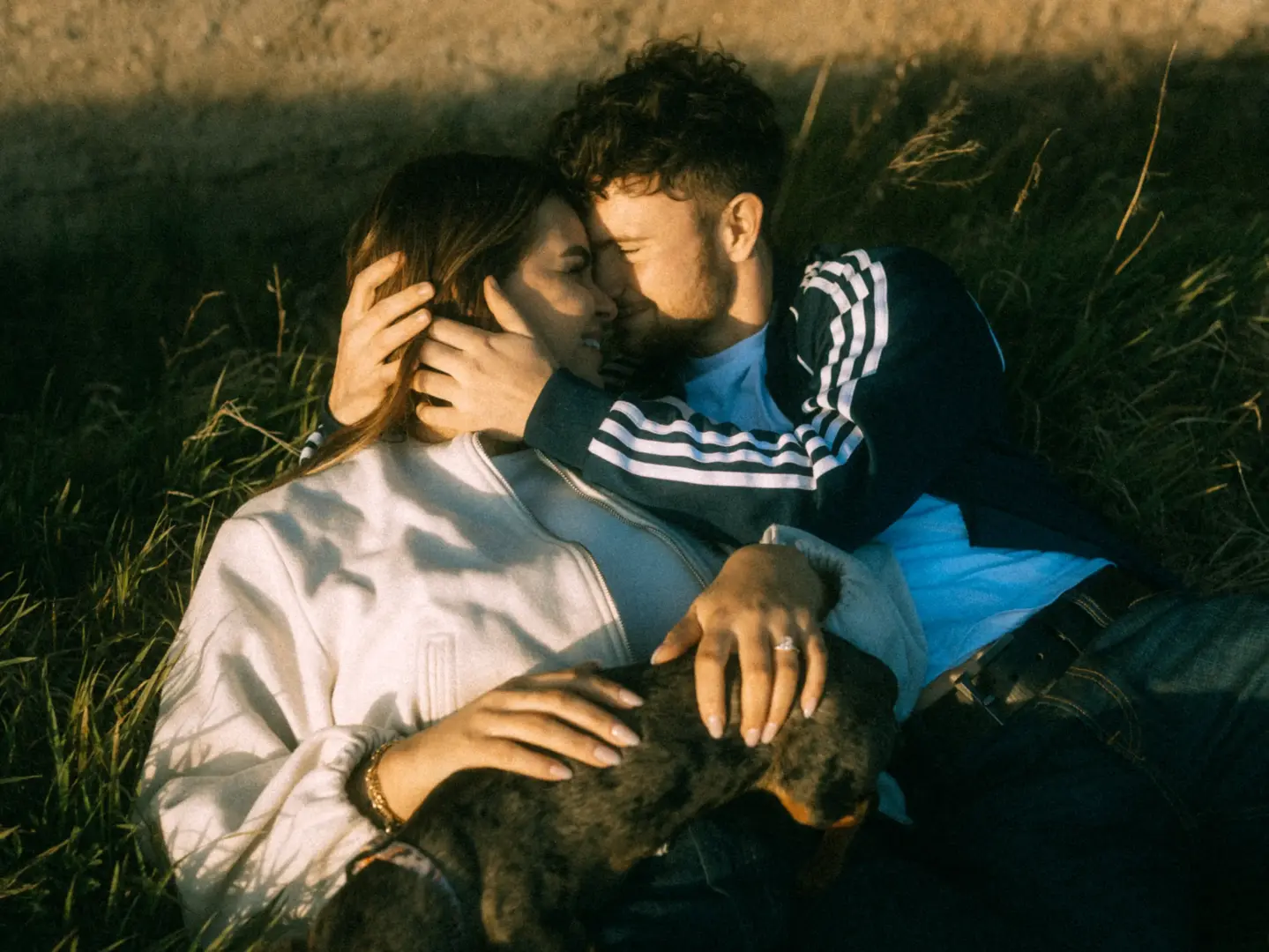 Ein junges Paar liegt eng umschlungen und verliebt im hohen Gras während des goldenen Sonnenuntergangs, während sie gemeinsam ihren kleinen Hund streicheln. Die romantische Atmosphäre unterstreicht eine professionelle Lovestory als Fotograf für Pärchen in Schwerin.